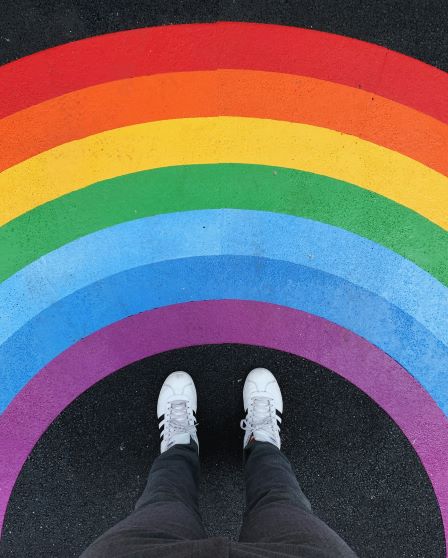 Photo of a rainbow painted onto asphalt. The photographer's shoes are visible from this angle